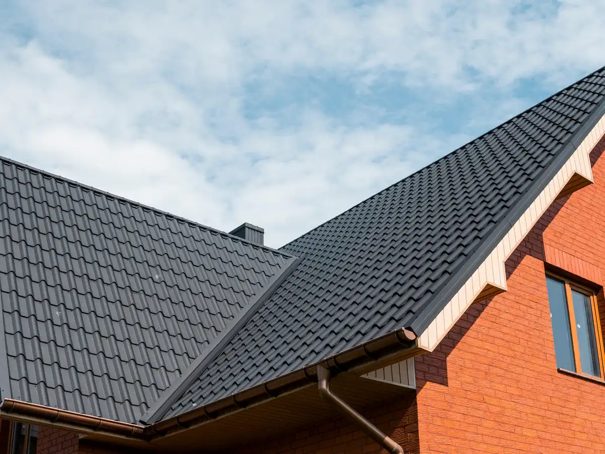 A close-up of a black wave-profile metal roofing system on a red brick house, showing the valley transition and brown guttering.