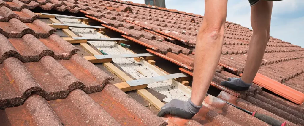 A roofing contractor wearing gloves inspecting wooden battens and removing terracotta tiles during a residential clay tile roof replacement.