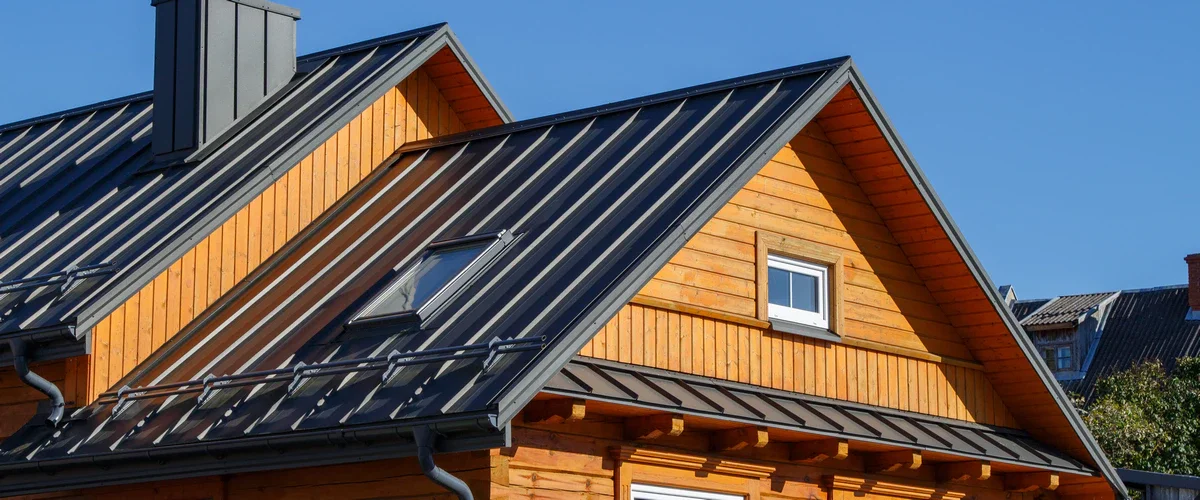 A dark gray standing seam metal roof installed on a rustic wooden log house featuring a skylight window and safety snow guards.