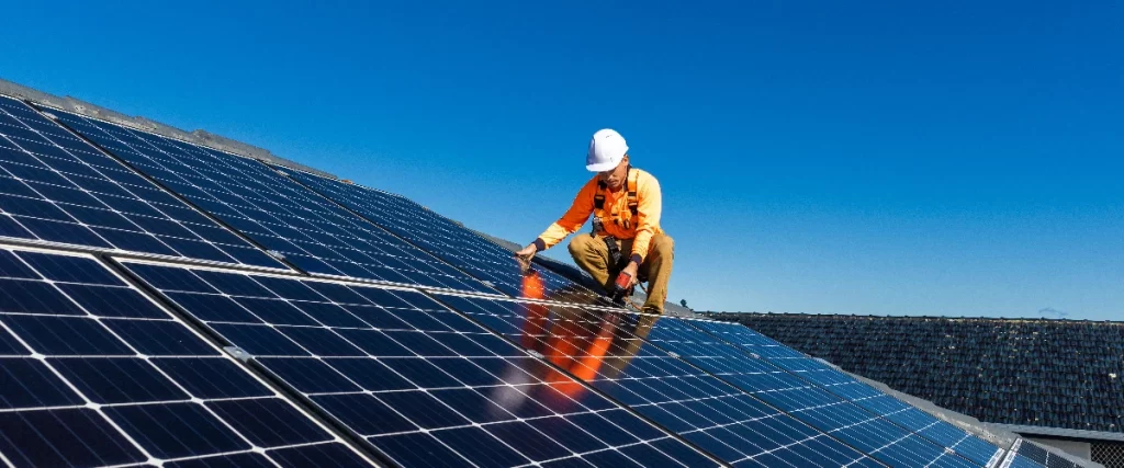 A male roofing contractor wearing an orange shirt, safety harness, and white hard hat uses a drill to secure a solar panel onto a dark residential roof under a clear blue sky.