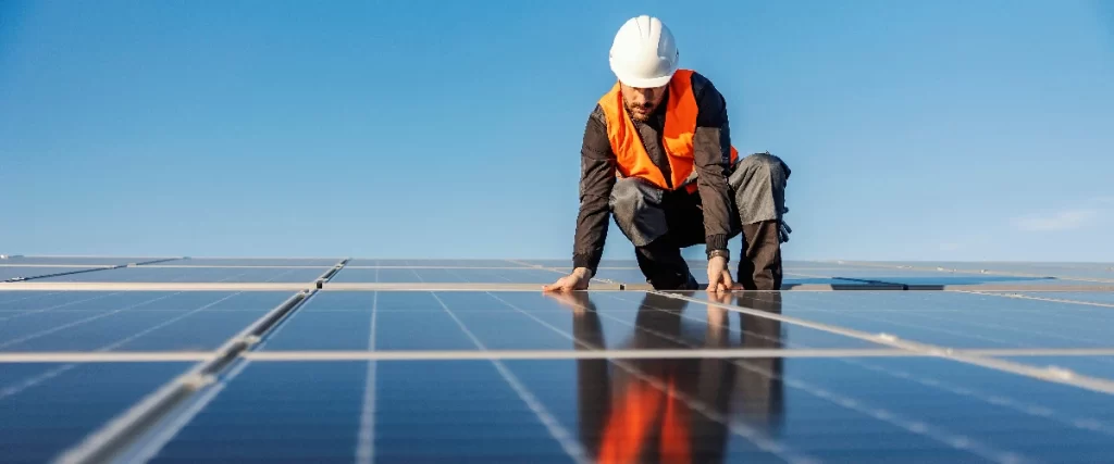 A male worker wearing an orange high-visibility vest and a white hard hat crouches down to carefully inspect a clean, reflective solar panel on a bright sunny day.