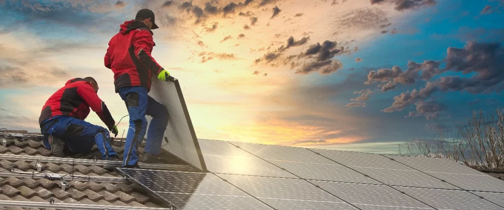 Two installation technicians wearing red and black jackets carefully lift and align a large solar panel onto a tiled roof mounting system against a dramatic sunset sky.