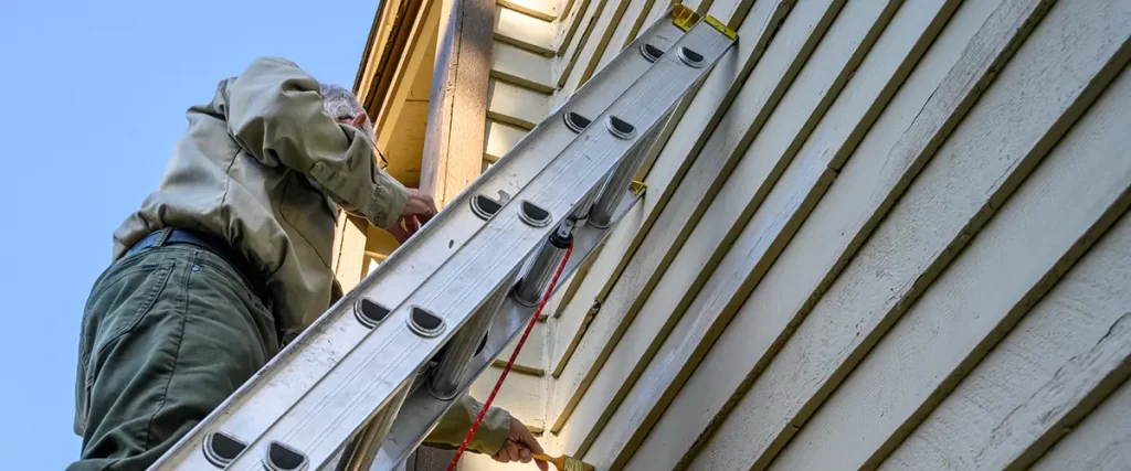 Homeowner painting exterior siding from a ladder as part of house siding maintenance and repair