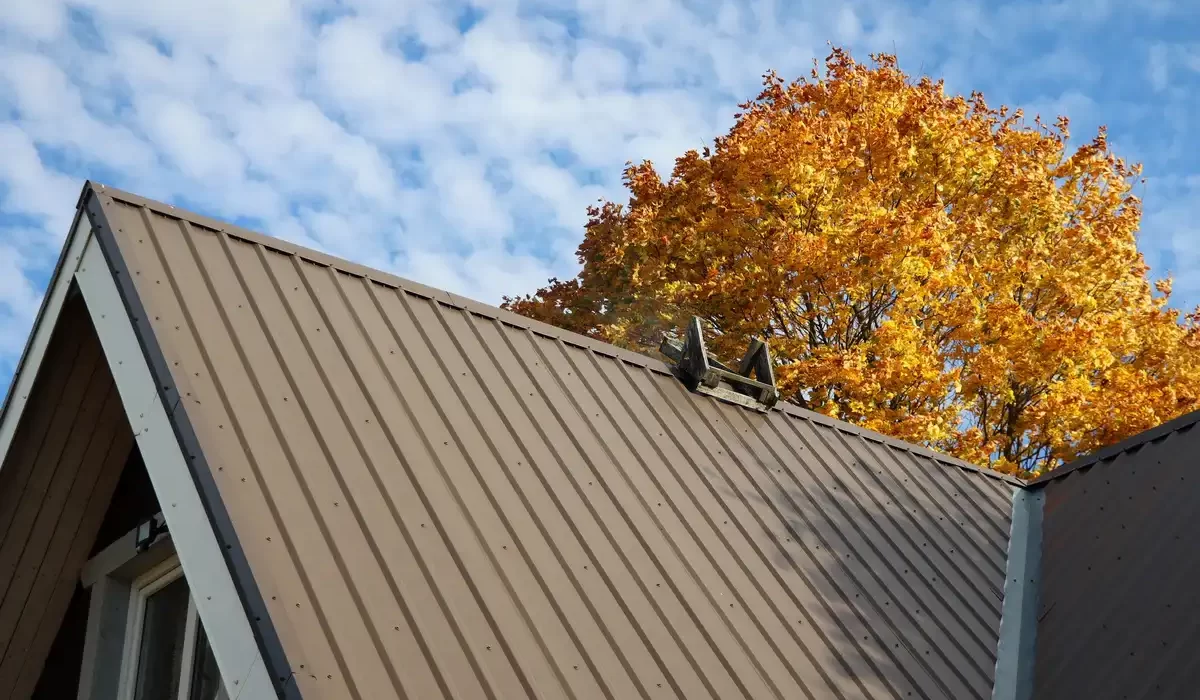 A tan ribbed panel metal roofing gable shown against a bright blue sky and a vibrant orange autumn tree in the background.